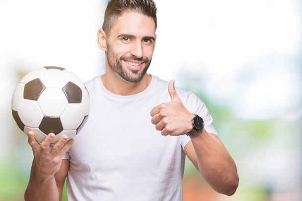 Νέος άνθρωπος που κρατά μπάλα ποδοσφαίρου ποδοσφαίρου πέρα από το απομονωμένο υπόβαθρο ευτυχισμένη με μεγάλο χαμόγελο κάνει εντάξει σήμα, τον αντίχειρα επάνω με τα δάχτυλα, καλό σημάδι - Φωτογραφία, εικόνα