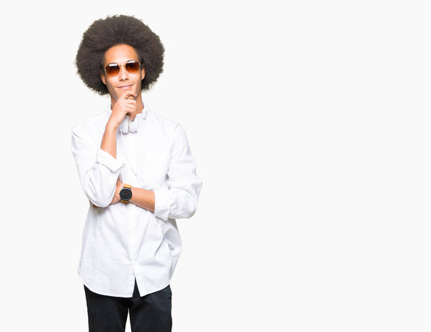 Jonge Afro-Amerikaanse man met afro haar dragen zonnebril en hoofdtelefoon met hand op kin denken over de vraag, nadenkend expressie. Glimlachend met doordachte gezicht. Twijfel concept. - Foto, afbeelding