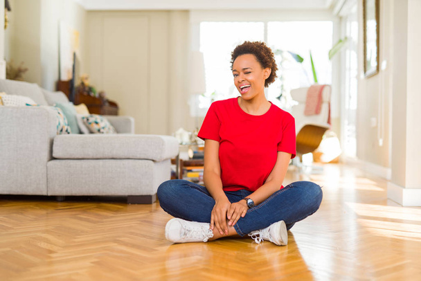 Jeune belle femme afro-américaine assise sur le sol à la maison clignant des yeux en regardant la caméra avec une expression sexy, gaie et heureuse visage
. - Photo, image