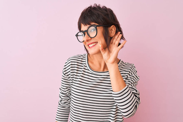 Молодая красивая женщина в полосатой футболке и в очках на изолированном розовом фоне улыбается рукой над ухом, слушая слухи или сплетни. Концепция глухоты
. - Фото, изображение