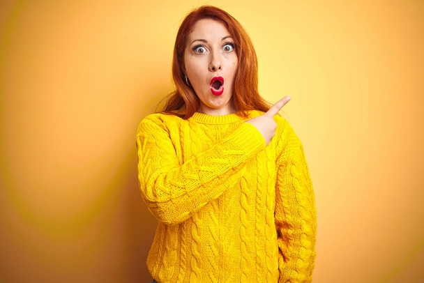 Belle femme rousse portant un pull d'hiver debout sur fond jaune isolé Surprise pointant du doigt sur le côté, bouche ouverte expression étonnante
. - Photo, image