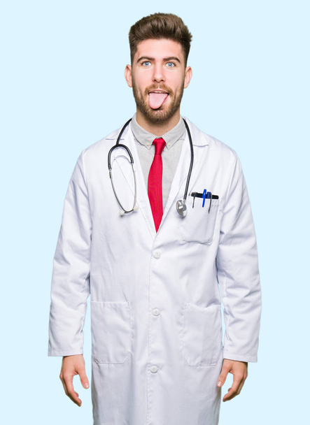 Young handsome doctor man wearing medical coat sticking tongue out happy with funny expression. Emotion concept. - Foto, immagini