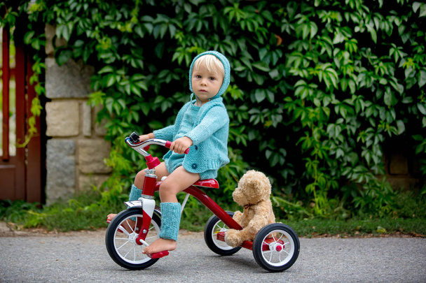 Adorable toddler boy with knitted outfit, riding tricycle on a q - Zdjęcie, obraz