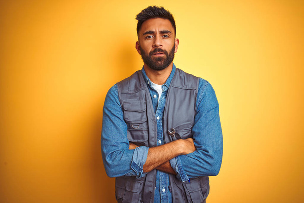 Handsome indian technician man wearing uniform over isolated yellow background skeptic and nervous, disapproving expression on face with crossed arms. Negative person. - Photo, image