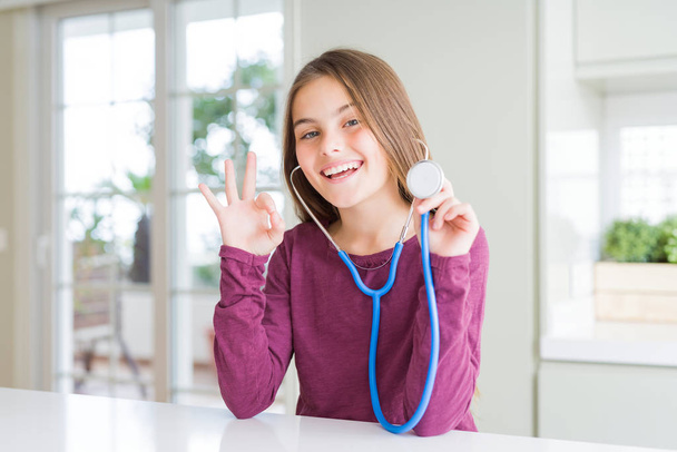 Kaunis nuori tyttö lapsi tarkistaa terveyttä käyttäen lääkärin stetoskooppi tekee ok merkki sormilla, erinomainen symboli
 - Valokuva, kuva