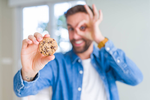 Bonito homem comendo biscoitos de chocolate com rosto feliz sorrindo fazendo ok sinal com a mão no olho olhando através dos dedos
 - Foto, Imagem