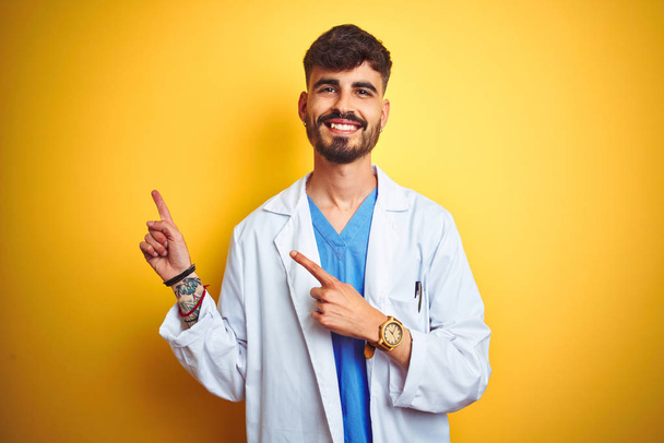 Nuori lääkäri, jolla on tatuointi, seisoo eristetyn keltaisen taustan päällä hymyillen ja katsoo kameraa osoittaen kahdella kädellä ja sormella sivulle
. - Valokuva, kuva