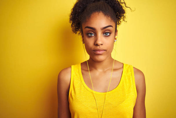African american woman listening to music using earphones over isolated yellow background with serious expression on face. Simple and natural looking at the camera. - 写真・画像
