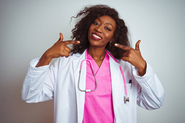 Africká americká doktorka, Žena s růžovým stetoskem nad osamělým bílým pozadím, usměvavá a ukazující na prsty zuby a ústa. Zdravotní koncepce. - Fotografie, Obrázek