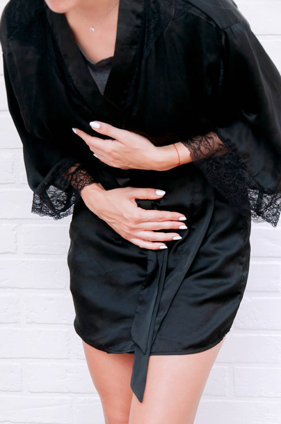 A girl holds on to her stomach in a black coat against a white wall. The girl has abdominal pain. - Foto, imagen