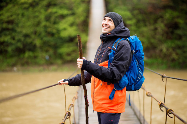 Man with backpack trekking in forest by hinged bridge over river. Cold weather. Autumn hiking. Wooden bridge across the river, Suspension bridge - Photo, Image