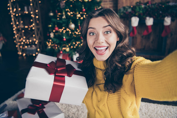 Self portrait of cheerful rejoicing girl shooting her gift box with christmas tree behind wearing yellow sweater - Фото, изображение
