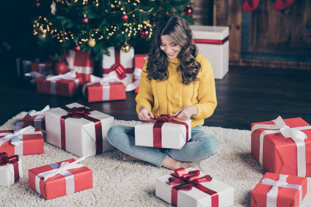 Photo of charming nice cute girl wearing jeans denim yellow sweater unpacking her new year gift from santa claus while sitting on carpet on floor with legs crossed - Фото, изображение