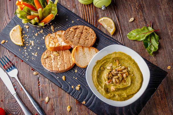 Hummus com espinafre, sementes de abacate e abóbora em uma tigela em uma tábua de madeira e bruschetta, cozinha oriental, orientação horizontal, vista superior
 - Foto, Imagem