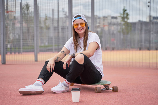 Image of smiling athletic brunette girl in sunglasses sitting on skateboard on playground on summer day - Foto, afbeelding
