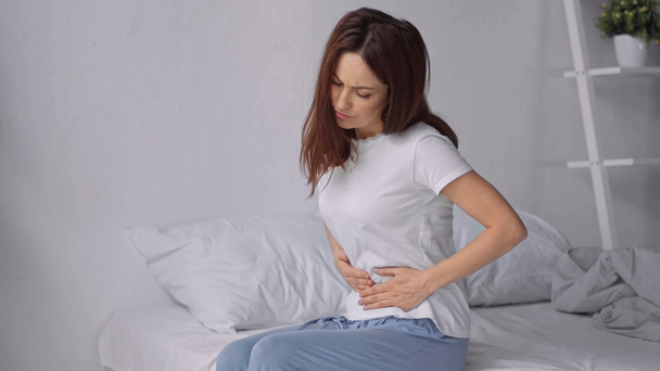 Frau sitzt mit Bauchschmerzen im Schlafanzug im Bett - Filmmaterial, Video