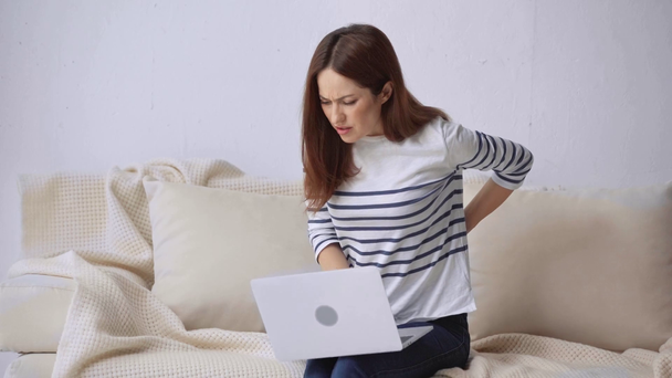mujer usando el ordenador portátil en el sofá y sufriendo de dolor de espalda
 - Metraje, vídeo