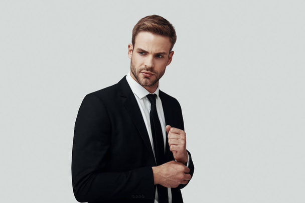 Handsome young man in formalwear looking away and adjusting sleeve while standing against grey background - Foto, Bild