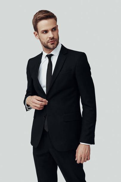 Bello giovane in abbigliamento formale guardando altrove e regolando giacca mentre in piedi su sfondo grigio
 - Foto, immagini