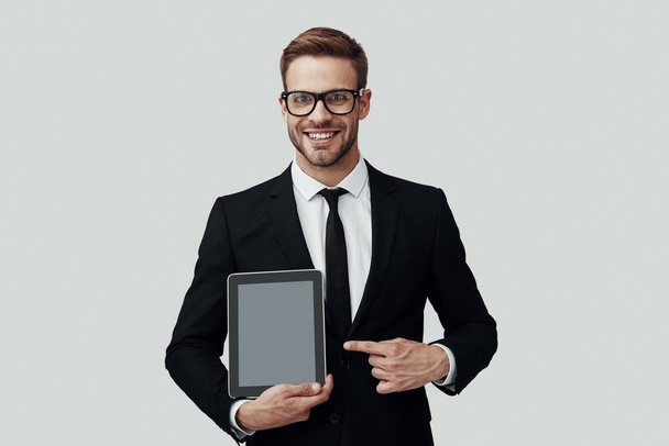Красивий молодий чоловік у формальному одязі, що показує простір копіювання на цифровому планшеті, стоячи на сірому фоні
 - Фото, зображення