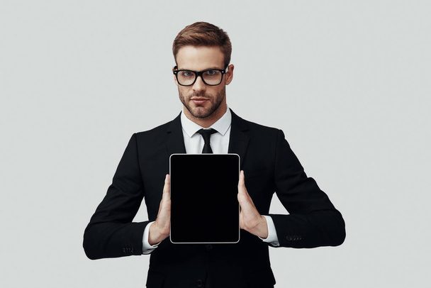 Przystojny młody człowiek w formalwear pokazując przestrzeń kopiowania na tablecie cyfrowym, stojąc na szarym tle - Zdjęcie, obraz