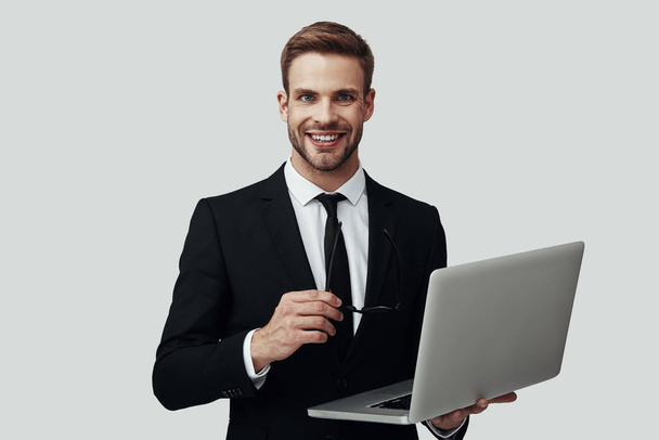 Красивый молодой человек в формальной одежде работает с ноутбуком и улыбается стоя на сером фоне
 - Фото, изображение