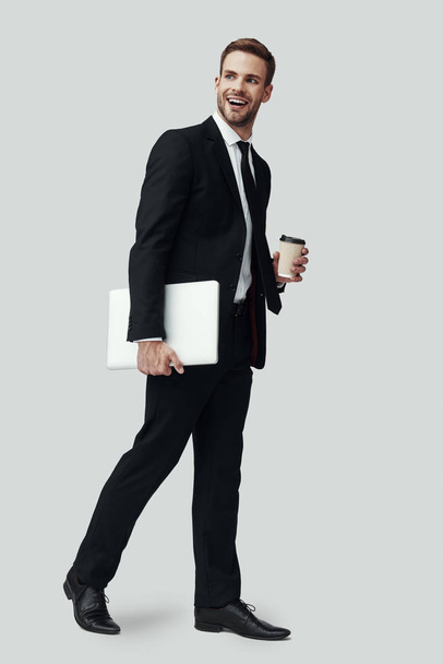 Piena lunghezza di bel giovane uomo in abbigliamento formale che trasporta il computer portatile e distogliendo lo sguardo mentre in piedi sullo sfondo grigio
 - Foto, immagini