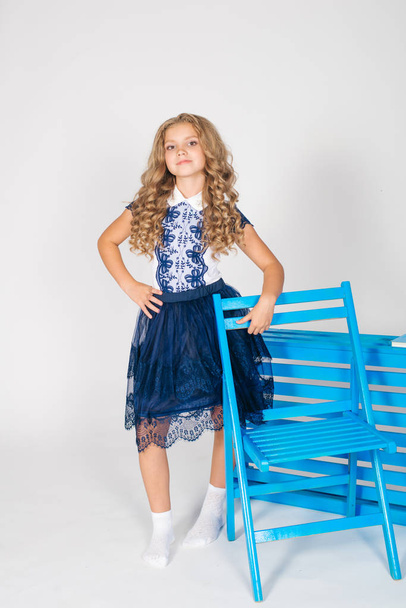 Cute girl with long blond curly hair in school fashion clothes with blue chair on a white background - Foto, afbeelding