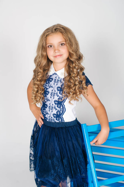 Cute girl with long blond curly hair in school fashion clothes with blue chair on a white background - Foto, immagini