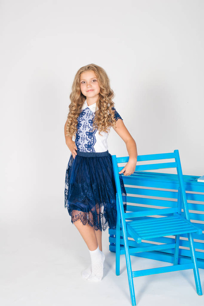 Cute girl with long blond curly hair in school fashion clothes with blue chair on a white background - Fotografie, Obrázek