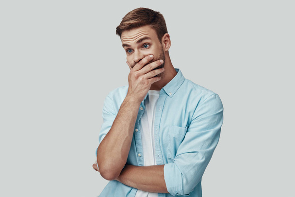 Surprised young man covering mouth with hand while standing against grey background - Fotoğraf, Görsel