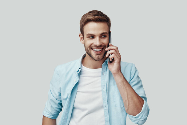 Pohledný mladý muž, který mluví po telefonu a usmívá se, když stojí proti šedému pozadí - Fotografie, Obrázek