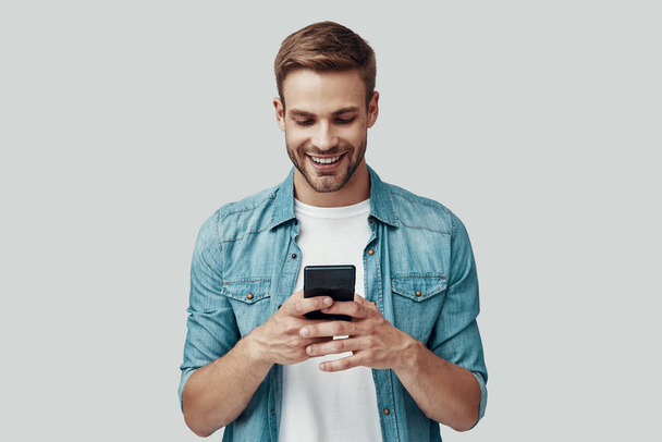 Pohledný mladý muž, který používá chytrý telefon a usmívá se, když stojí proti šedému pozadí - Fotografie, Obrázek