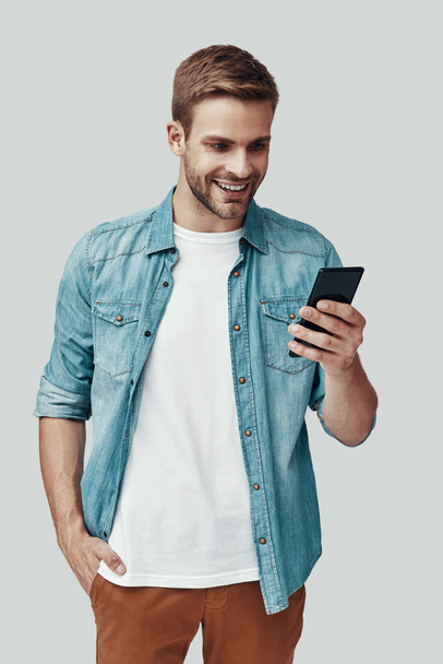 schöner junger Mann mit Smartphone und lächelnd vor grauem Hintergrund - Foto, Bild