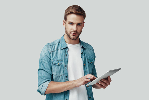 Csinos fiatalember nézi a kamerát, és digitális tabletta használata közben állva szürke háttér - Fotó, kép