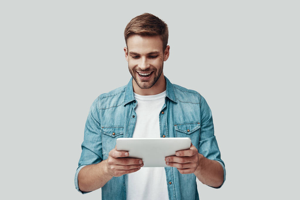 Handsome young man smiling and using digital tablet while standing against grey background - Фото, изображение