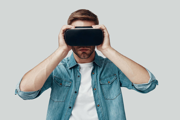 Handsome young man using virtual reality simulator while standing against grey background - Photo, Image