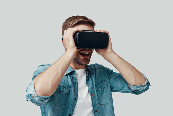schöner junger Mann im Virtual-Reality-Simulator und lächelnd vor grauem Hintergrund - Foto, Bild