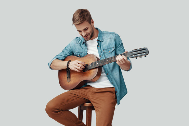 schöner junger Mann lächelt und spielt Gitarre, während er vor grauem Hintergrund sitzt - Foto, Bild