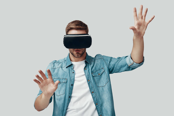 Όμορφος νέος άνθρωπος χρησιμοποιώντας προσομοιωτή εικονική πραγματικότητα, ενώ στέκεται ενάντια σε γκρίζο φόντο - Φωτογραφία, εικόνα