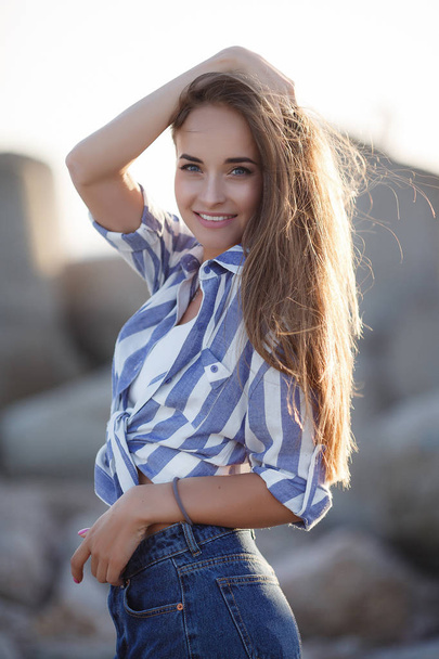 Beautiful brunette sitting on the rocks.Sexy brunette girl posing alone on the beach in the evening.Young pretty hot sexy attractive girl relaxing in in a striped shirt and shorts on stones with blue sea and sky on background.Summer Vacation Concept. - 写真・画像