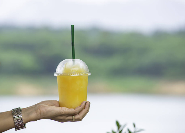 Kädessä lasi kylmää appelsiinimehua smoothie Tausta hämärä näkymät puu ja vesi
. - Valokuva, kuva