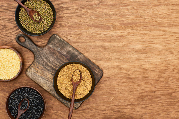 top view of cutting board with bowls with black beans, maash, couscous and buckwheat on wooden surface - Photo, Image