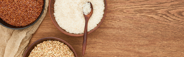 panoramic shot of bowls with white rice with spoon near roasted and raw buckwheat on wooden surface with canvas - Photo, Image