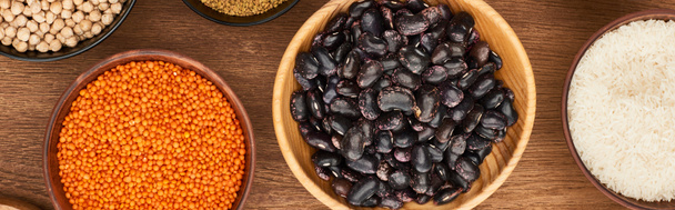 panoramic shot of bowls with red lentil, black beans, white rice and chickpea on wooden surface - Photo, Image