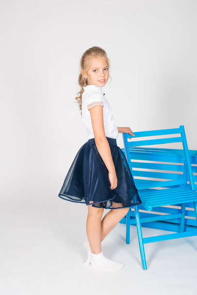 Cute girl with long blond curly hair in school fashion clothes with blue chair on a white background - Valokuva, kuva