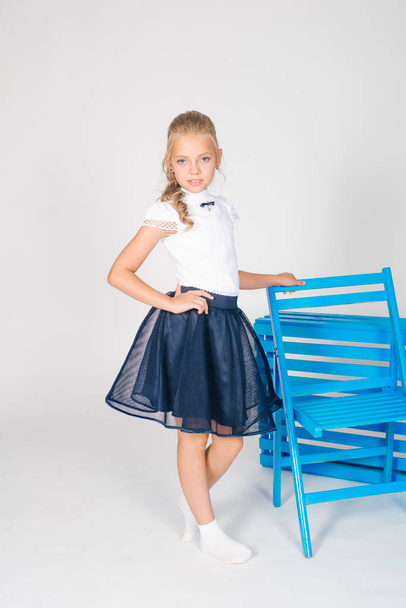 Cute girl with long blond curly hair in school fashion clothes with blue chair on a white background - Foto, Bild