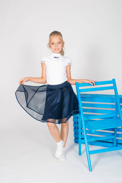 Cute girl with long blond curly hair in school fashion clothes with blue chair on a white background - Zdjęcie, obraz