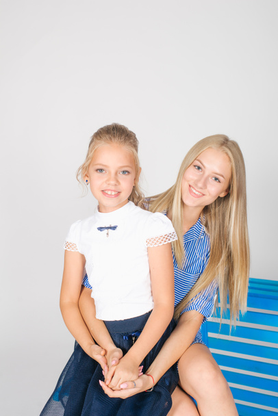 Roztomilá dívka s dlouhými blonďatými vlasy ve školních módních šatech s modrou židlí na bílém pozadí s mladým učitelem s mladým učitelem - Fotografie, Obrázek