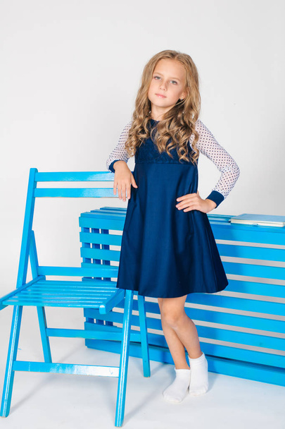 Cute girl with  blond curly hair in school fashion clothes with blue chair on a white background - Zdjęcie, obraz